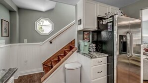 A kitchen with a stainless steel refrigerator, white cabinetry, a wooden staircase, and a window with blinds. - Ultimate vacay home, walk to beach, fenced in yard (North Myrtle Beach)