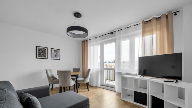 Un salon moderne avec un canapé gris, une table à manger avec des chaises, un téléviseur posé sur un meuble blanc et de grandes fenêtres avec des rideaux.