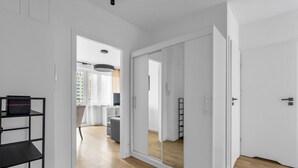 Un intérieur moderne avec un plancher en bois, des murs blancs et un grand miroir. On y trouve une étagère en métal noir et une porte qui mène à une autre pièce.