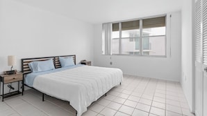 A bedroom with a bed, nightstands, a lamp, and a window with blinds. - Bay Harbor Sunset Apt 4B (Bay Harbor Islands)