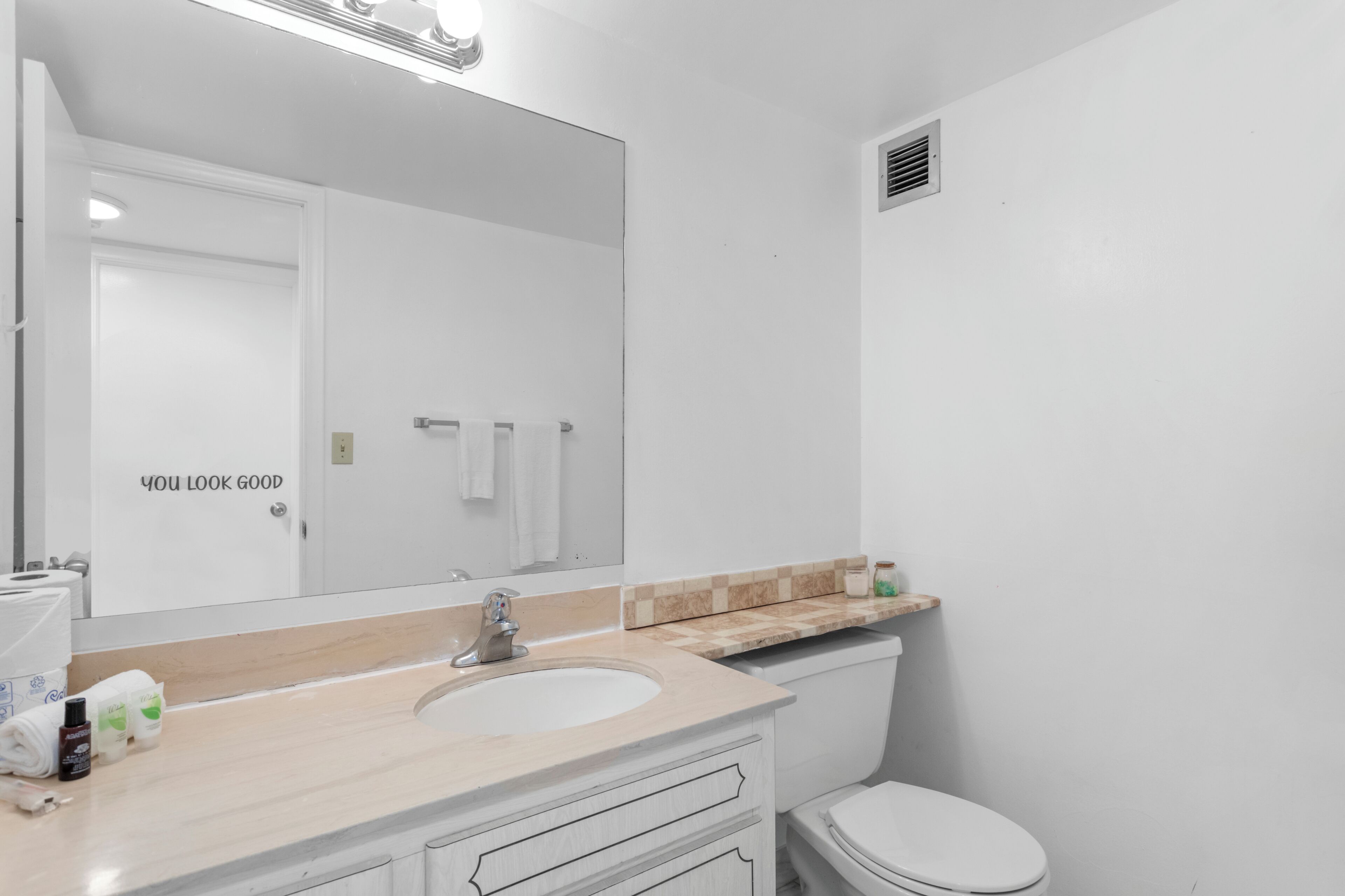 Ein Badezimmer mit weißem Waschbecken, Spiegel und Toilette. Darüber befindet sich ein Handtuchhalter und darüber eine kleine Ablagefläche mit Pflegeprodukten.