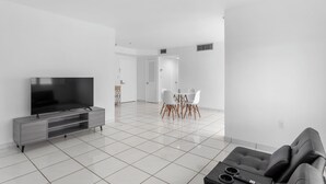 A modern living room with a flat-screen TV, grey sofa, and a dining area with white chairs and a table. - Bay Harbor Sunset Apt 4B (Bay Harbor Islands)