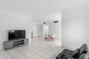 A modern living room with a flat-screen TV, grey sofa, and a dining area with white chairs and a table.