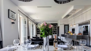 Ein moderner Essbereich mit einem langen Tisch, der für eine Mahlzeit gedeckt ist, einer Vase mit Blumen und einer Küche im Hintergrund.