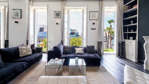 Ein geräumiges Wohnzimmer mit einem schwarzen Sofa, einem weißen Ottomanen und einem Glastisch. Offene Türen führen auf einen Balkon mit Blick auf Palmen und ein blaues Gebäude.