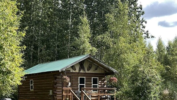 Un chalet rustique en rondins de bois, doté d’un porche, entouré d’une pelouse verdoyante et d’une forêt dense.