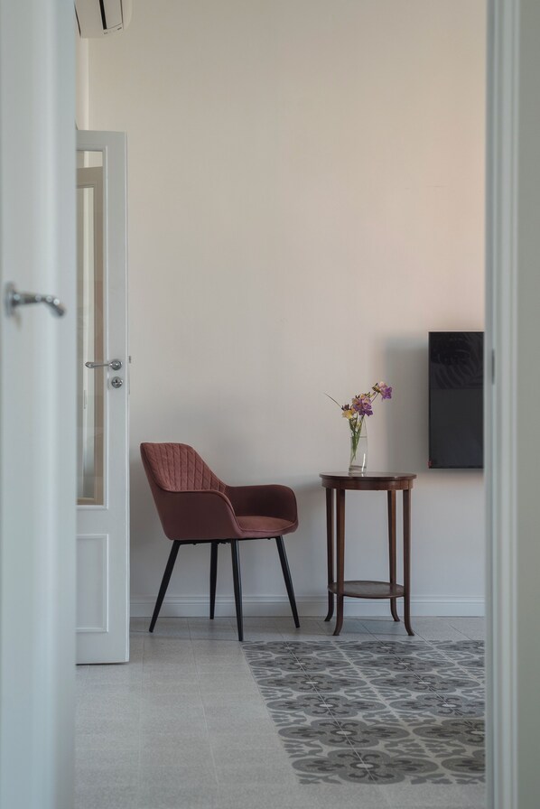 A room with a door, a chair, a small table with a vase of flowers, and a wall-mounted TV.