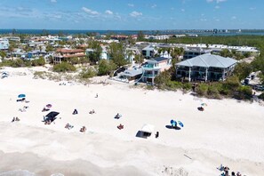 A coastal area with beach umbrellas, a large building with a balcony overlooking the beach, and a distant view of the ocean and a bridge.