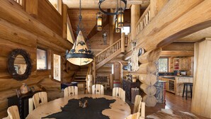 A log cabin interior with a round wooden dining table, a black leather centerpiece, wooden chairs, a staircase, and a kitchen area visible through an open door. - New! Ski-in/out Log Home beside 104.3 acres with a private hot tub (Big Sky)