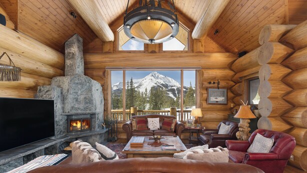 A log cabin interior with a fireplace, leather furniture, and a mountain view. - New! Ski-in/out Log Home beside 104.3 acres with a private hot tub (Big Sky)