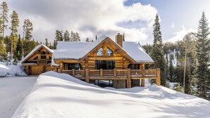 A log cabin with a snow-covered roof, wooden exterior, and a balcony. - New! Ski-in/out Log Home beside 104.3 acres with a private hot tub (Big Sky)