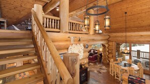 A log cabin interior with a wooden staircase, a dining area with a wooden table and chairs, and a cozy fireplace. - New! Ski-in/out Log Home beside 104.3 acres with a private hot tub (Big Sky)