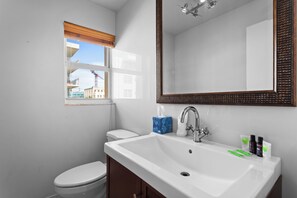 A bathroom with a white sink, a mirror, a window with a view of buildings, and a toilet. - Bay Harbor Sunset Apt 5A (Bay Harbor Islands)