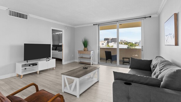 A modern living room with a flat-screen TV, a grey sofa, a wooden coffee table, and a sliding glass door leading to a balcony with a city view. - Bay Harbor Sunset Apt 5A (Bay Harbor Islands)