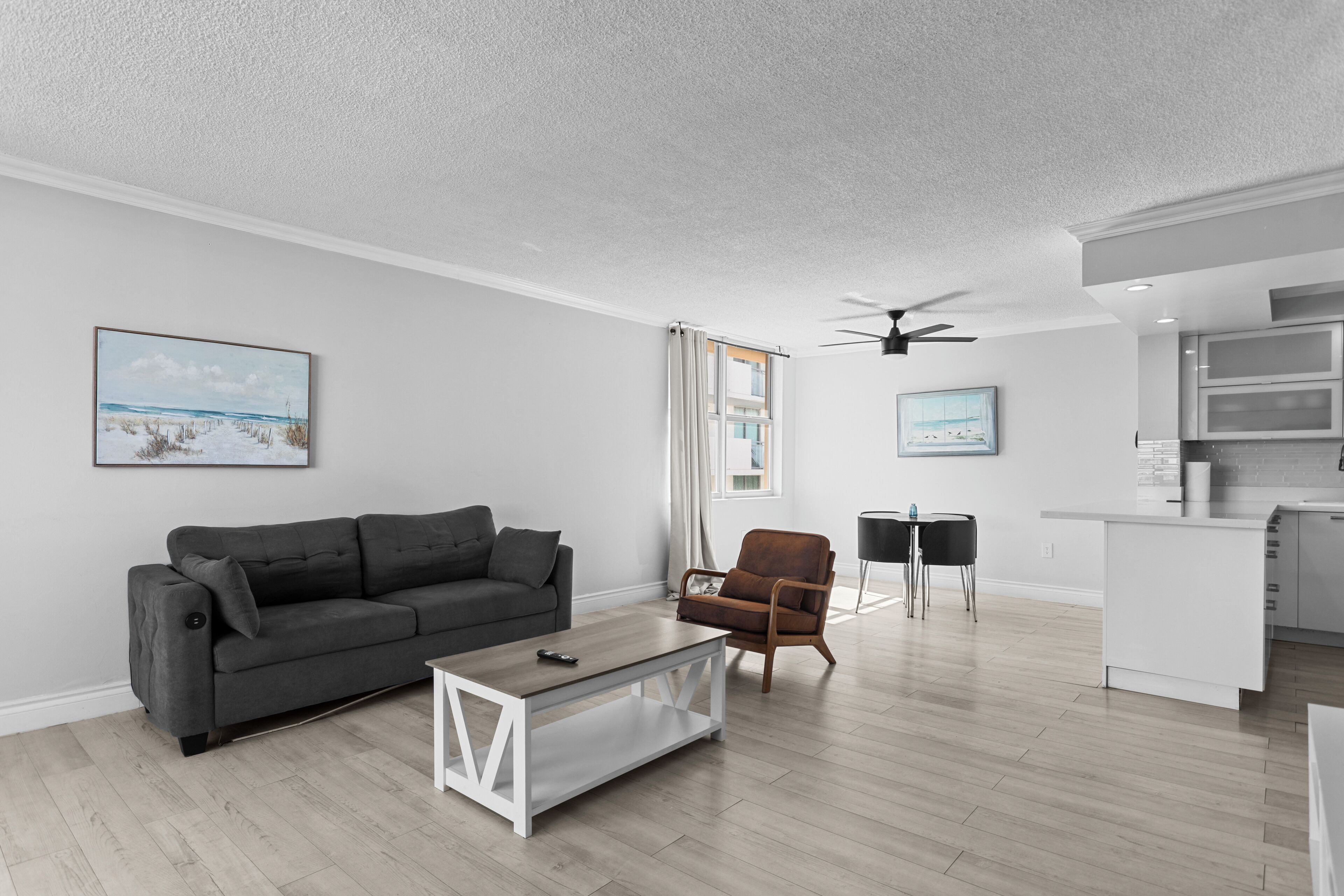 Un salon moderne avec un canapé gris, une table basse et un fauteuil marron. On y trouve également un coin cuisine avec un îlot central blanc et des placards.