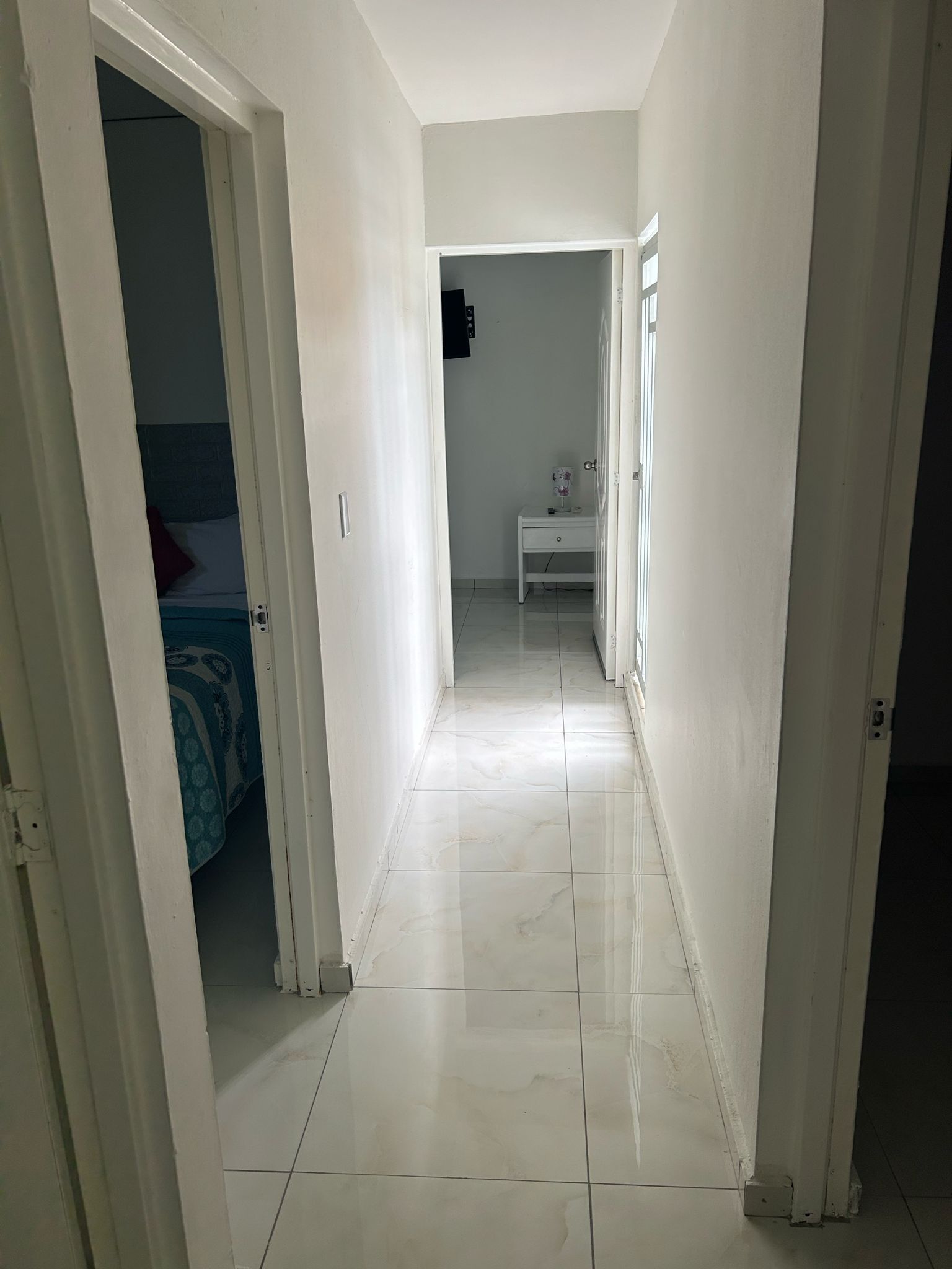 Un estrecho pasillo con paredes blancas y suelo de baldosas que conduce a un dormitorio con cama y mesita de noche.