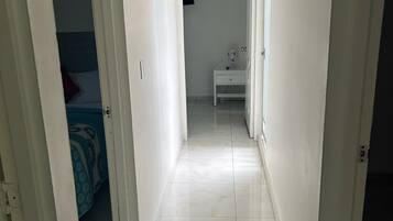 Un estrecho pasillo con paredes blancas y suelo de baldosas que conduce a un dormitorio con cama y mesita de noche.