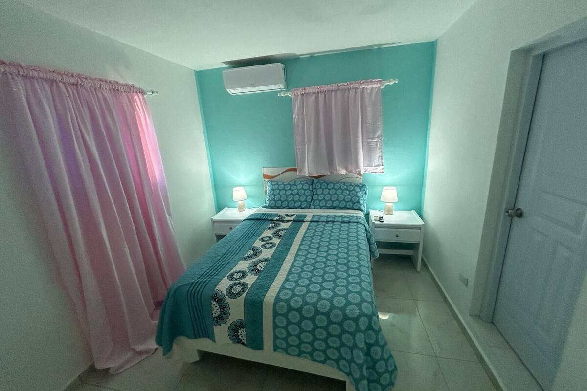 Un dormitorio con una cama, dos mesitas de noche, una ventana con cortinas y un equipo de aire acondicionado.