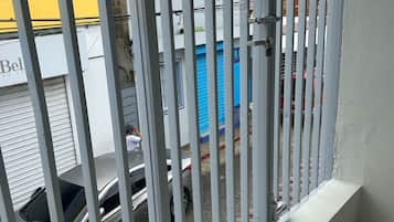 Un balcón con barandilla metálica que da a una calle con coches aparcados y un edificio con toldo amarillo.