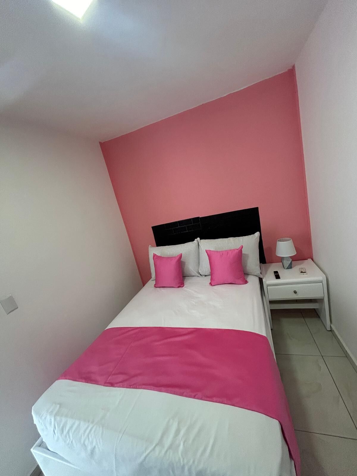 A bedroom with a bed, two pillows, a nightstand, a lamp, and a power outlet.