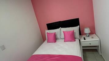 A bedroom with a bed, two pillows, a nightstand, a lamp, and a power outlet.
