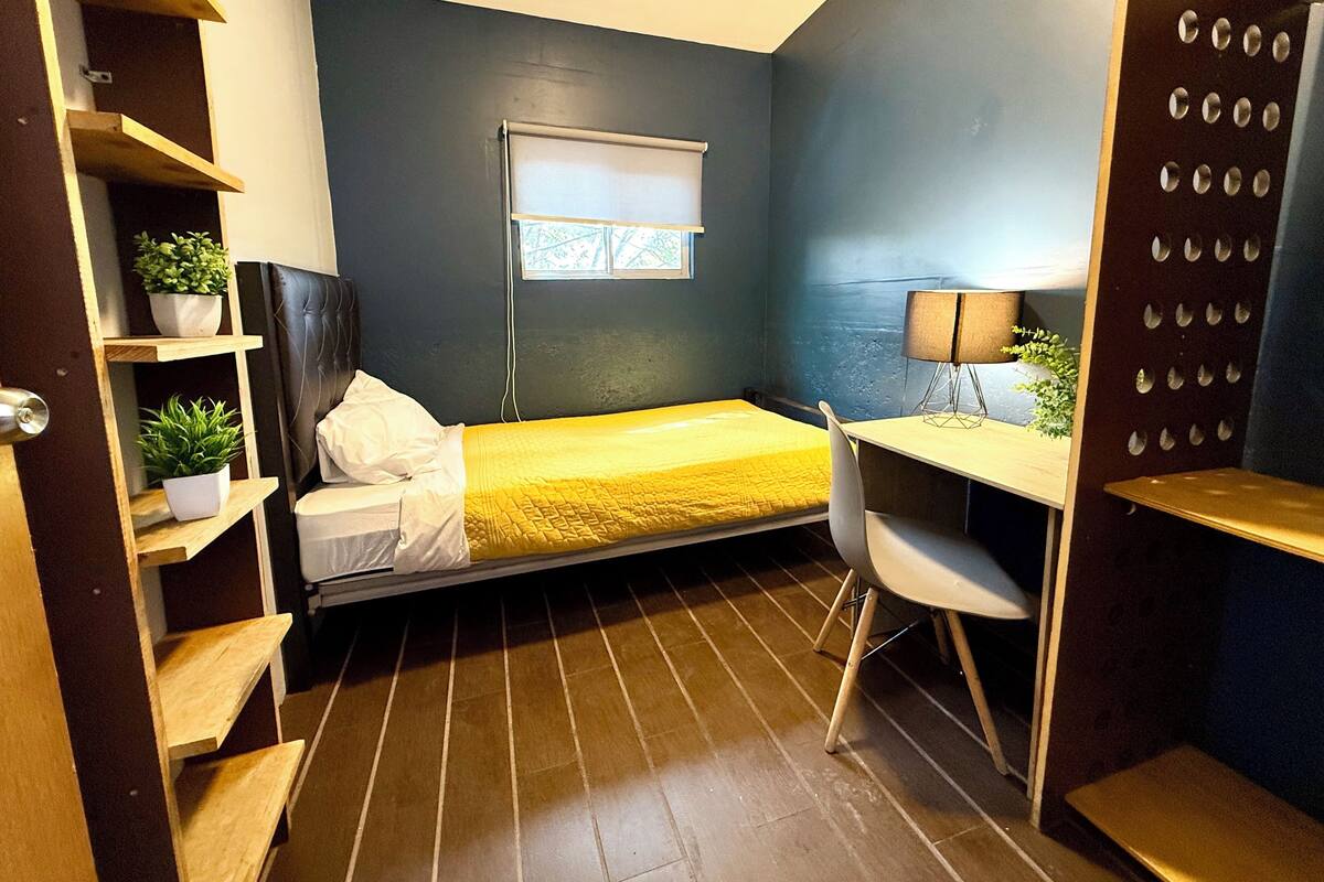 Ein kleines Zimmer mit einem Bett, einem Schreibtisch und Regalen mit Pflanzen.
