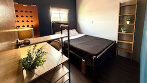 A compact room with a bed, a desk, a chair, a window, and a shelving unit. - WONDER HOSTAL - Santa Maria (Mexico City)