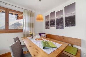 Een eetgedeelte met een houten tafel, stoelen, een bank, een hangende hanglamp en foto’s van een berglandschap aan de muur.