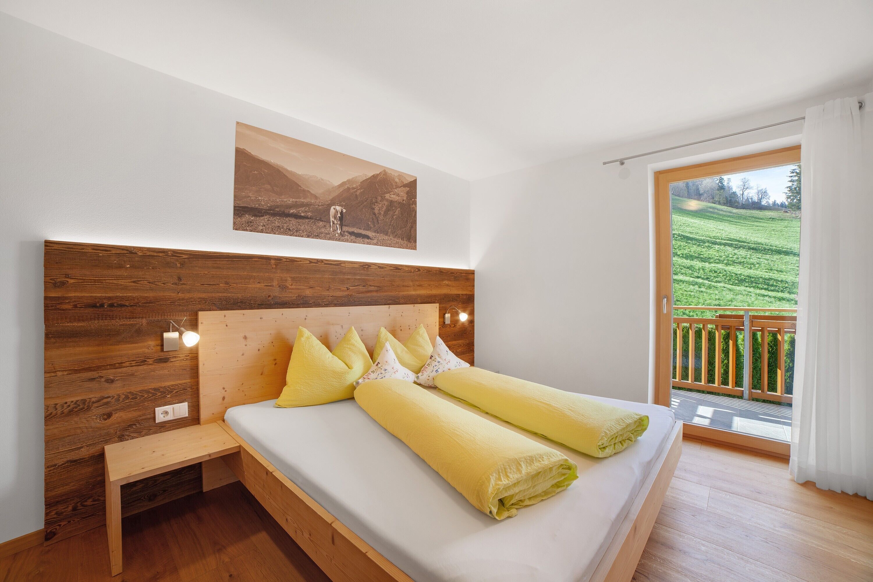 Une chambre à coucher avec une tête de lit en bois, un lit recouvert de draps jaunes, une table de chevet et une vue sur un paysage verdoyant par une grande fenêtre.