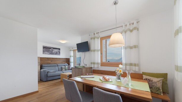A modern room with a wooden dining table, grey chairs, a sofa, a flat-screen TV, and a window with curtains. - Apartment 'Thymian' with Mountain View, Private Terrace and Wi-Fi (Schenna)