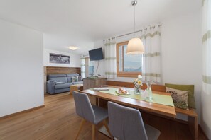 Een moderne kamer met een houten eettafel, grijze stoelen, een bank, een flatscreen televisie en een raam met gordijnen.