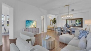 Een moderne woonkamer met een grote flatscreen televisie, een eethoek met een kroonluchter en een zithoek met banken en een salontafel.