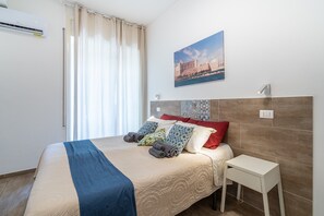 A neatly made bed with a blue blanket, a white bedside table, and a wall-mounted air conditioner.