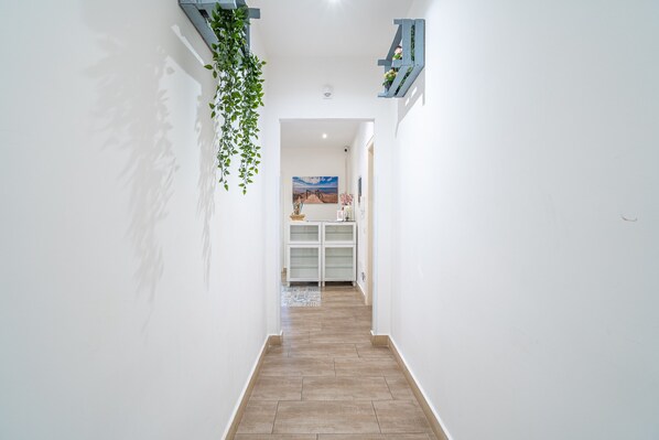 A hallway with a wooden floor, white walls, a hanging plant, a blue shelf, and a painting in the background.