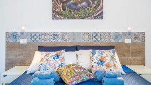 Una cama con un cojín estampado, una manta azul y vista a una escena costera.