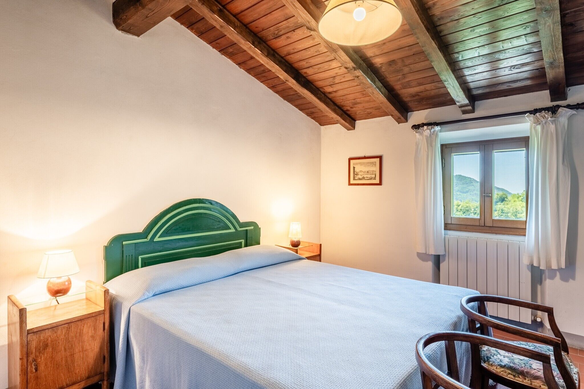 Une chambre avec un plafond en bois, un lit doté d’une tête de lit verte, une table de chevet avec une lampe, une chaise et une fenêtre avec des rideaux.