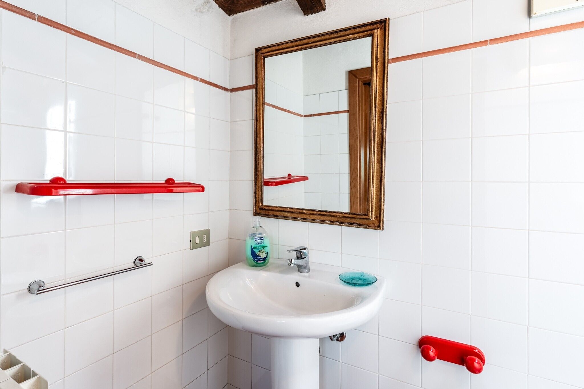 Une salle de bain avec des murs recouverts de carreaux blancs, un lavabo blanc, un miroir avec un cadre en bois et des porte-serviettes rouges.