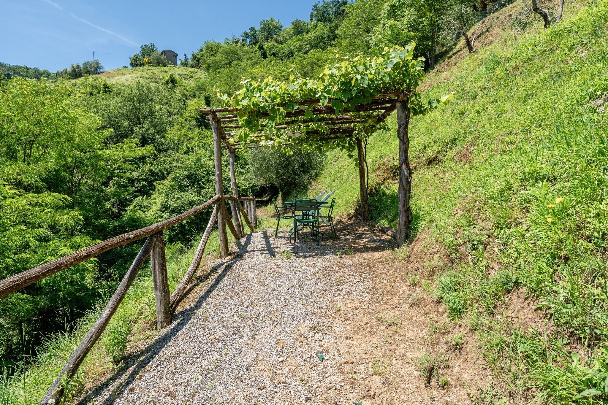 A gravel path leading to a wooden pergola with a table and chairs, surrounded by lush greenery.