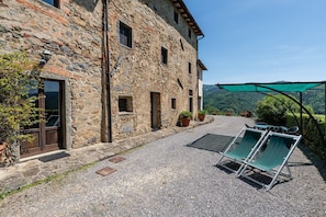 Un bâtiment en pierre avec une cour en gravier, des sièges à l’extérieur et une vue sur les collines.