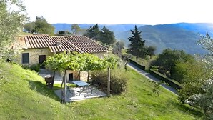 A stone house with a tiled roof, a small patio with a table and chairs, and a view of distant mountains. - Apartment 'Le Stalle' with Mountain View, Shared Pool and Wi-Fi (Pietrabuona)