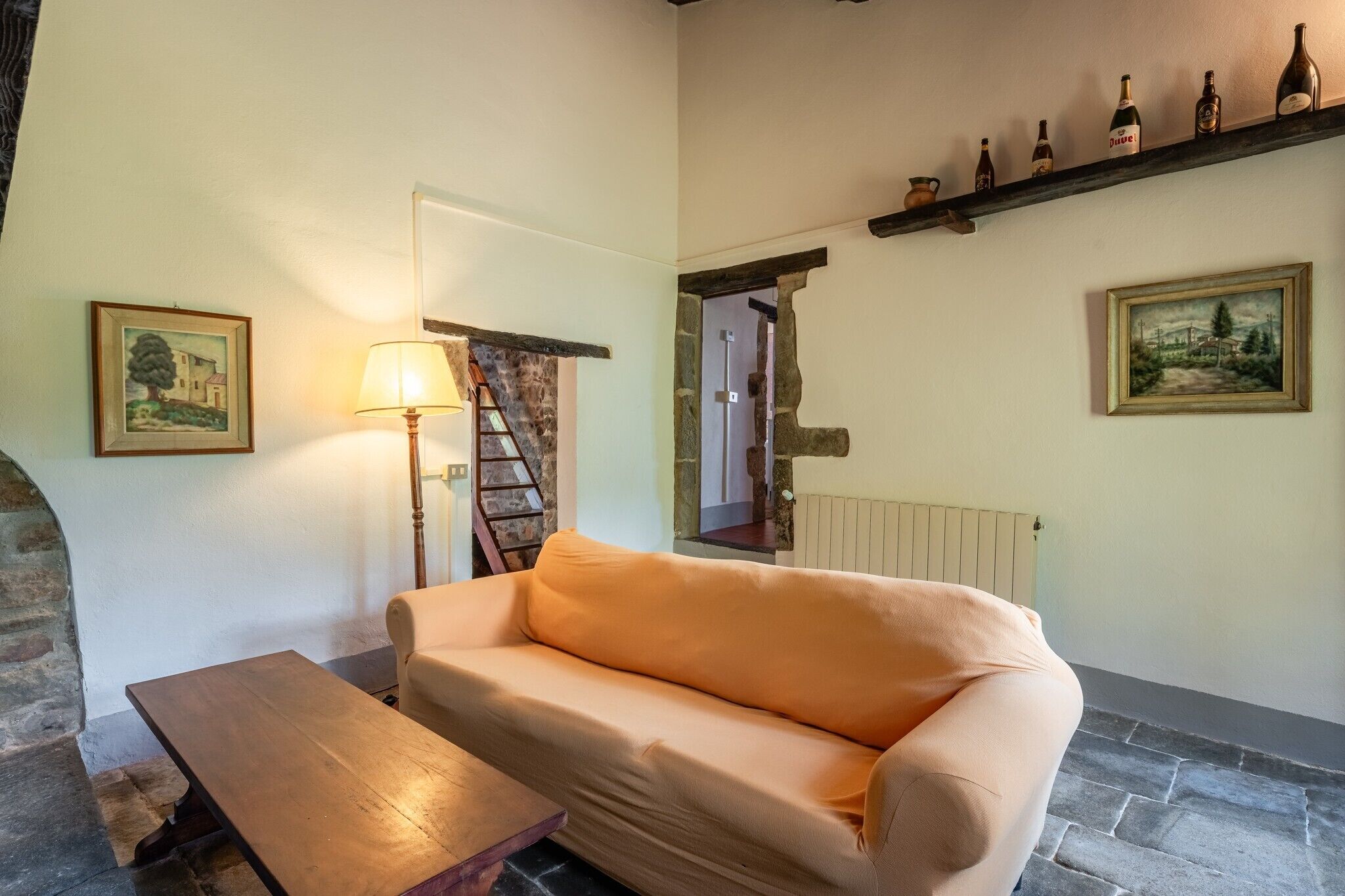 A cozy room with a sofa, a wooden table, a lamp, a ladder, a wine rack, and two paintings on the walls.