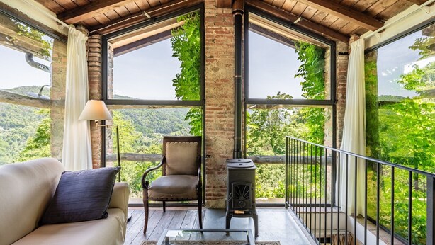 Une pièce chaleureuse avec un plafond en bois, de grandes fenêtres, un canapé, un fauteuil et un poêle à bois.