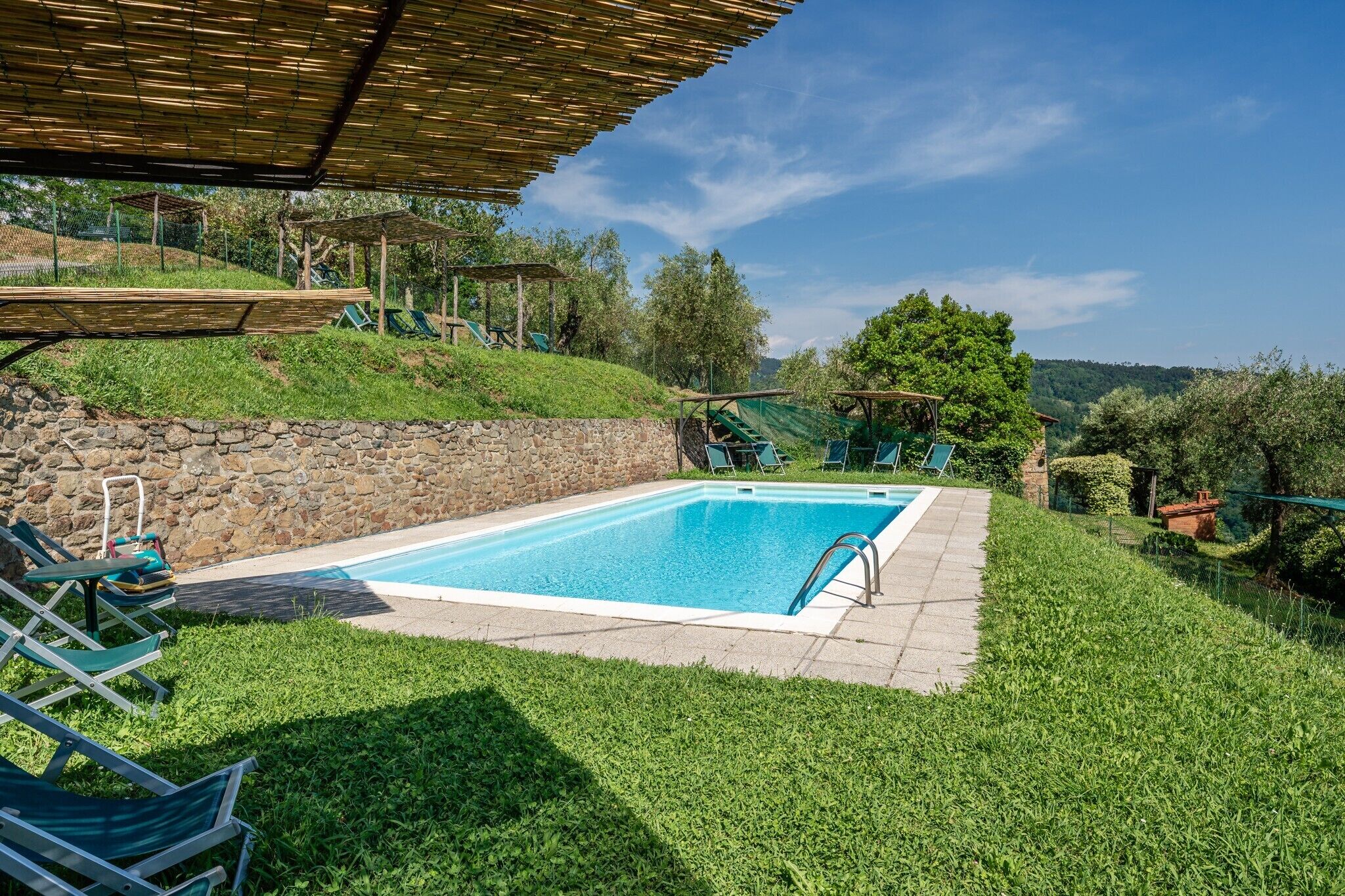 A swimming pool with a stone wall, green lawn, and a covered area with chairs.
