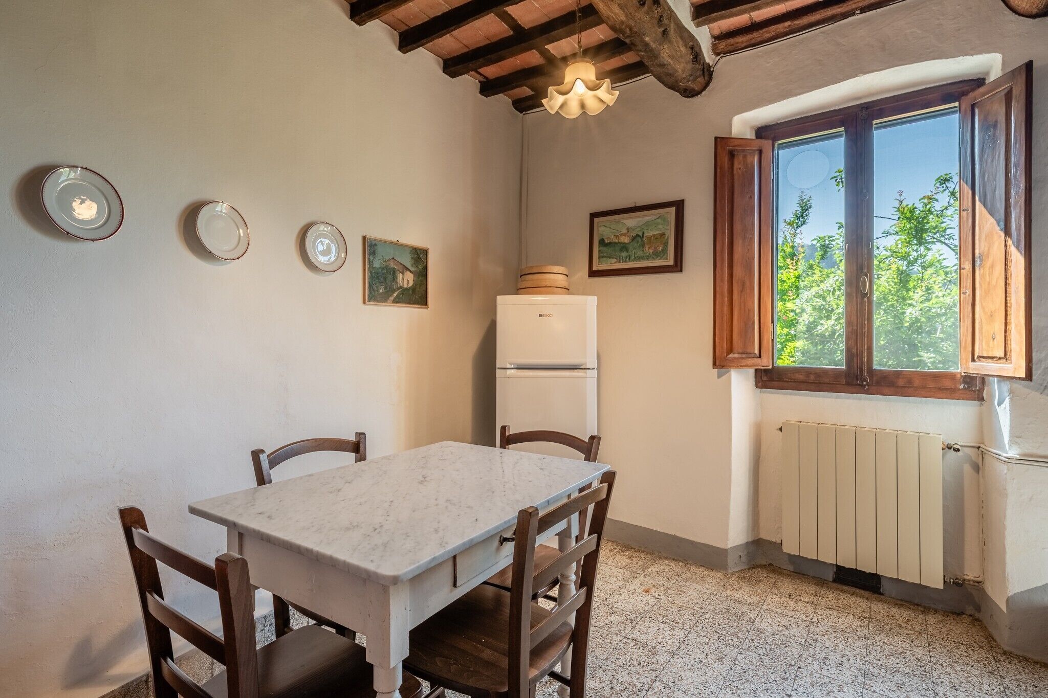 A dining area with a marble table, wooden chairs, a white refrigerator, and a window with a view of greenery.