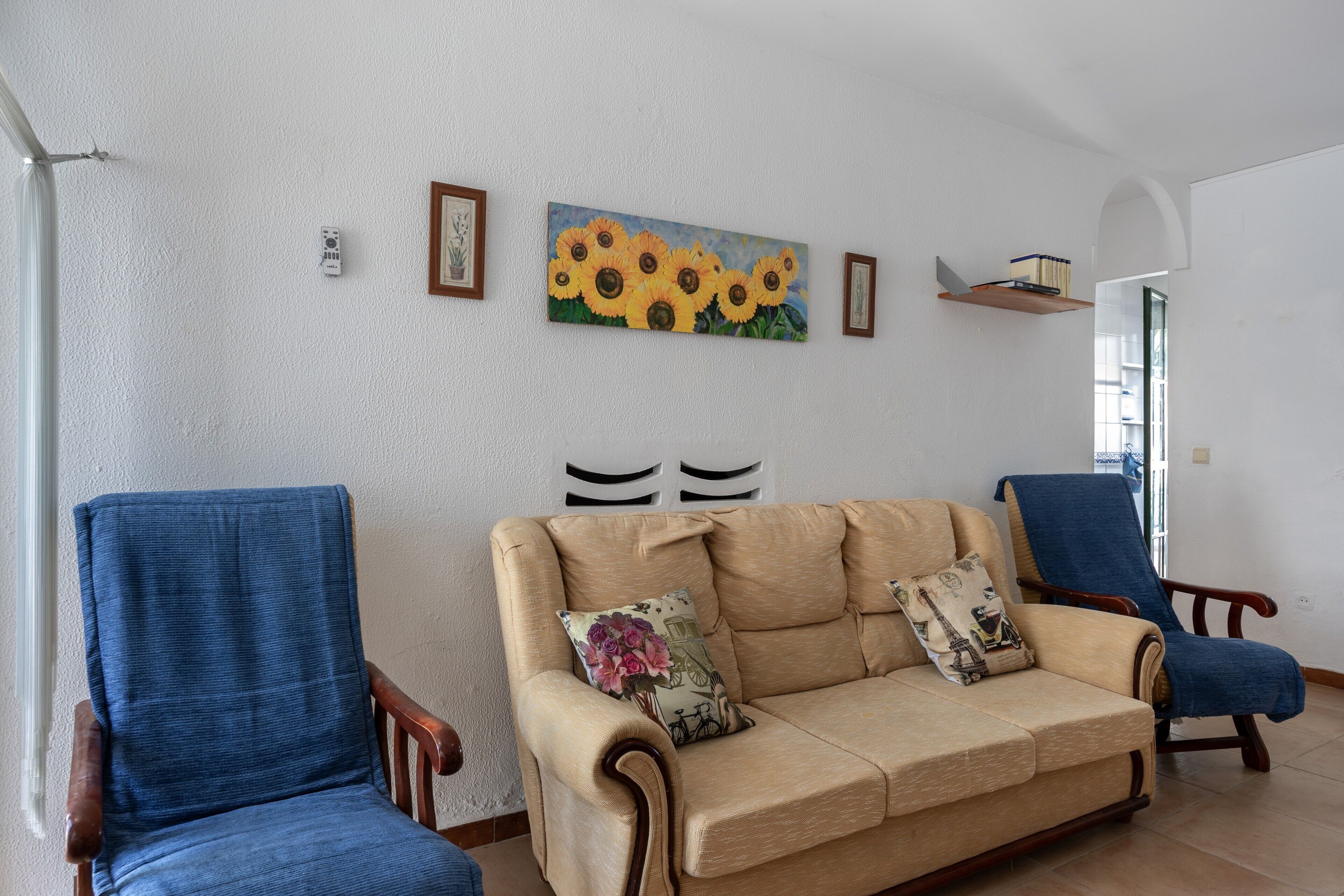Un salón con un sofá color beige, dos sillones azules y un cuadro de girasoles en la pared.
