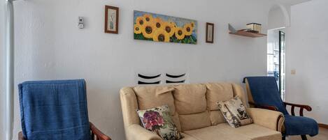 Un salón con un sofá color beige, dos sillones azules y un cuadro de girasoles en la pared.