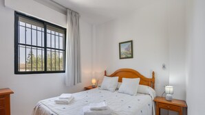 Une chambre à coucher avec un lit, deux tables de chevet, une fenêtre avec des barreaux, un tableau au mur et une vue sur l’extérieur.
