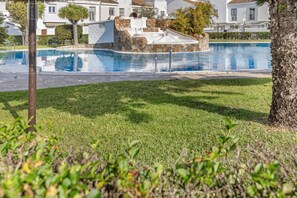 Una piscina con una estructura de piedra, rodeada de césped y palmeras, con edificios al fondo.
