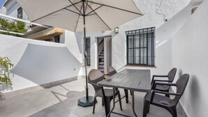 Une terrasse extérieure avec une table et des chaises, un grand parasol et un bâtiment blanc doté d’une fenêtre.