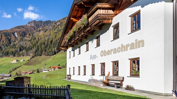 A white building with 'App. Oberachrain' signage, wooden balconies, and a view of a mountainous landscape.